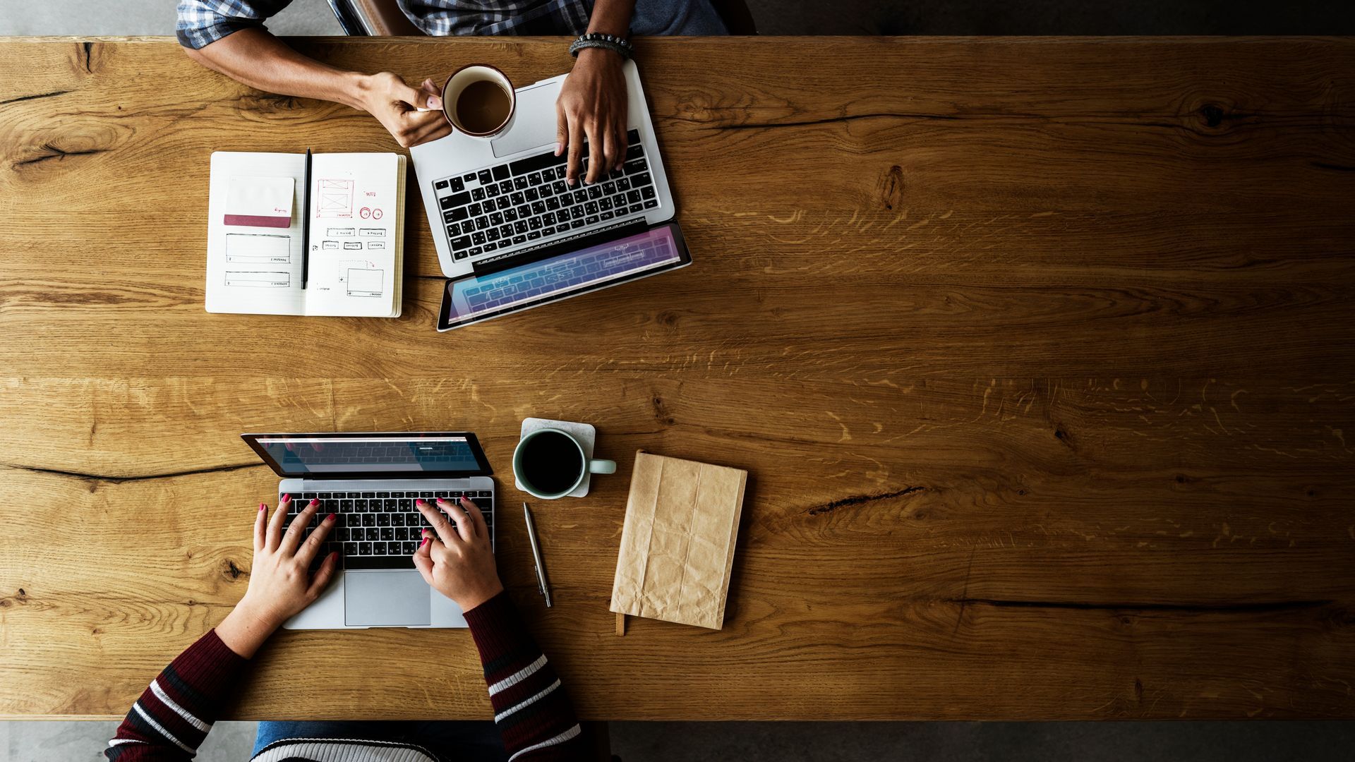 Deux personnes utilisent des ordinateurs portables à une table en bois, chacune avec un café et des notes. Vue de dessus.