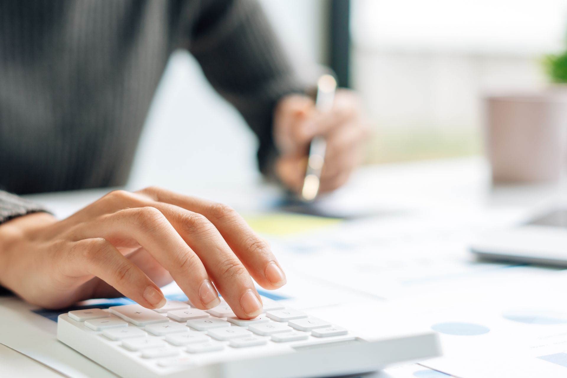 Les mains d'une personne utilisent une calculatrice blanche, une autre main tient un stylo au-dessus de feuilles de papier dont le fond est flou.