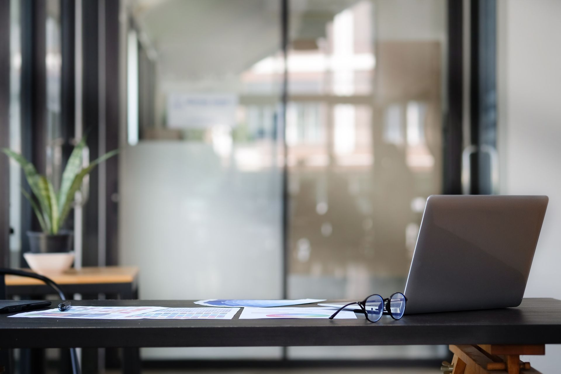 Un ordinateur portable posé sur un bureau, avec des lunettes, des papiers et une plante, dans un espace de bureau lumineux.