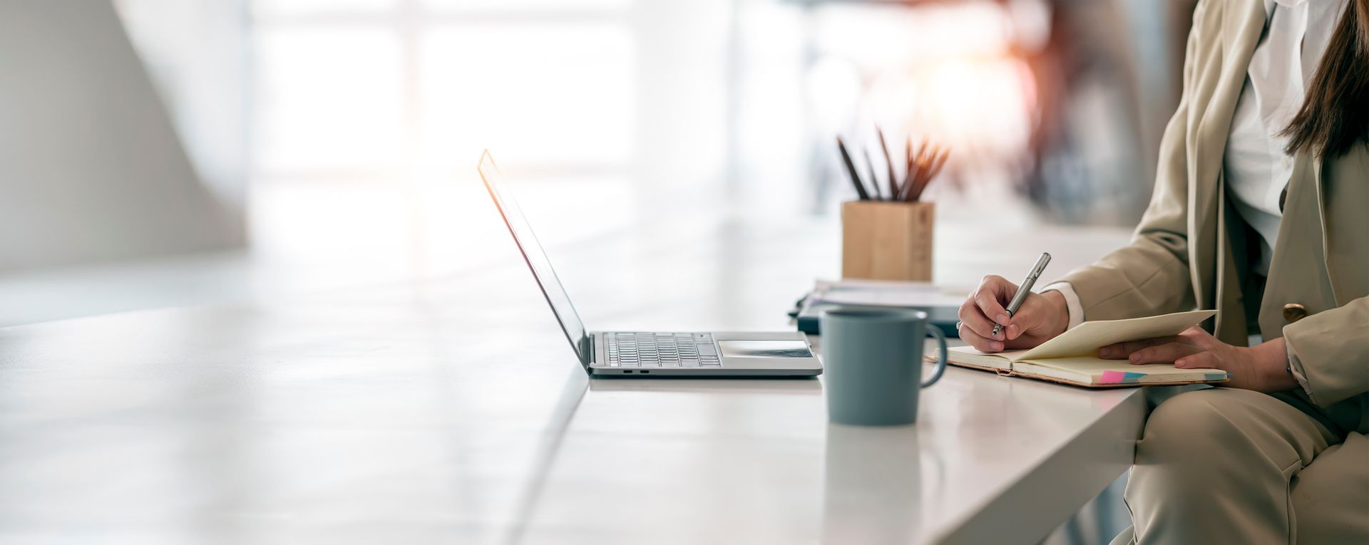 Une personne en costume beige prend des notes à un bureau avec un ordinateur portable, une tasse et des stylos.
