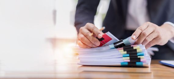 Une personne en costume trie une pile de documents sur un bureau, à l'aide d'un crayon et de pinces à reliure.