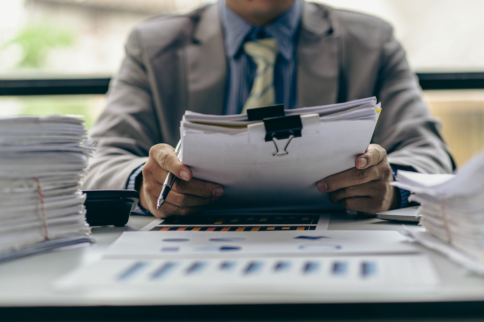 Une personne en costume examine des documents, un stylo à la main, au milieu de piles de papiers et de graphiques.