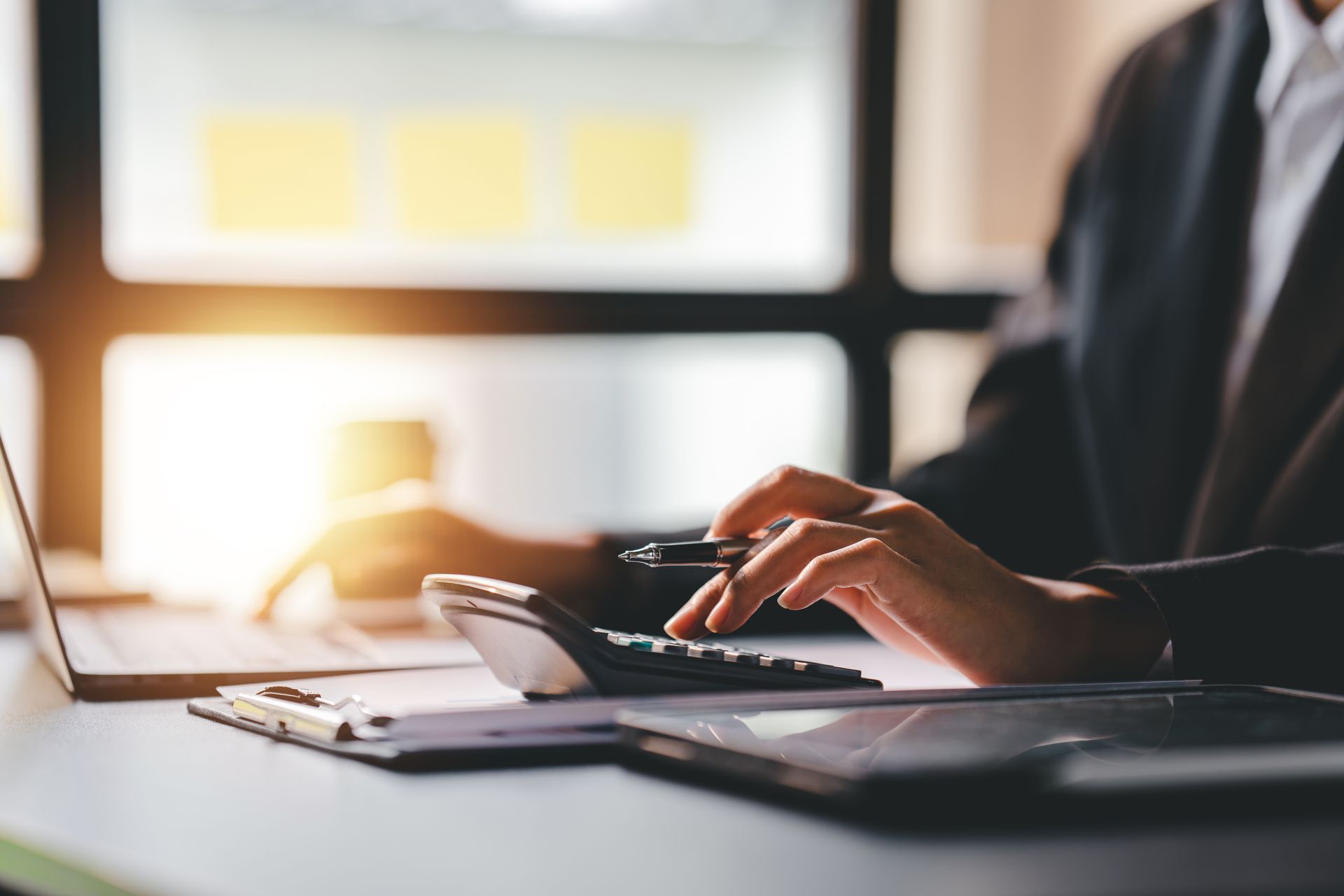 Une personne en costume utilise une calculatrice et un ordinateur portable à un bureau entouré de documents, éclairé par la lumière du soleil.