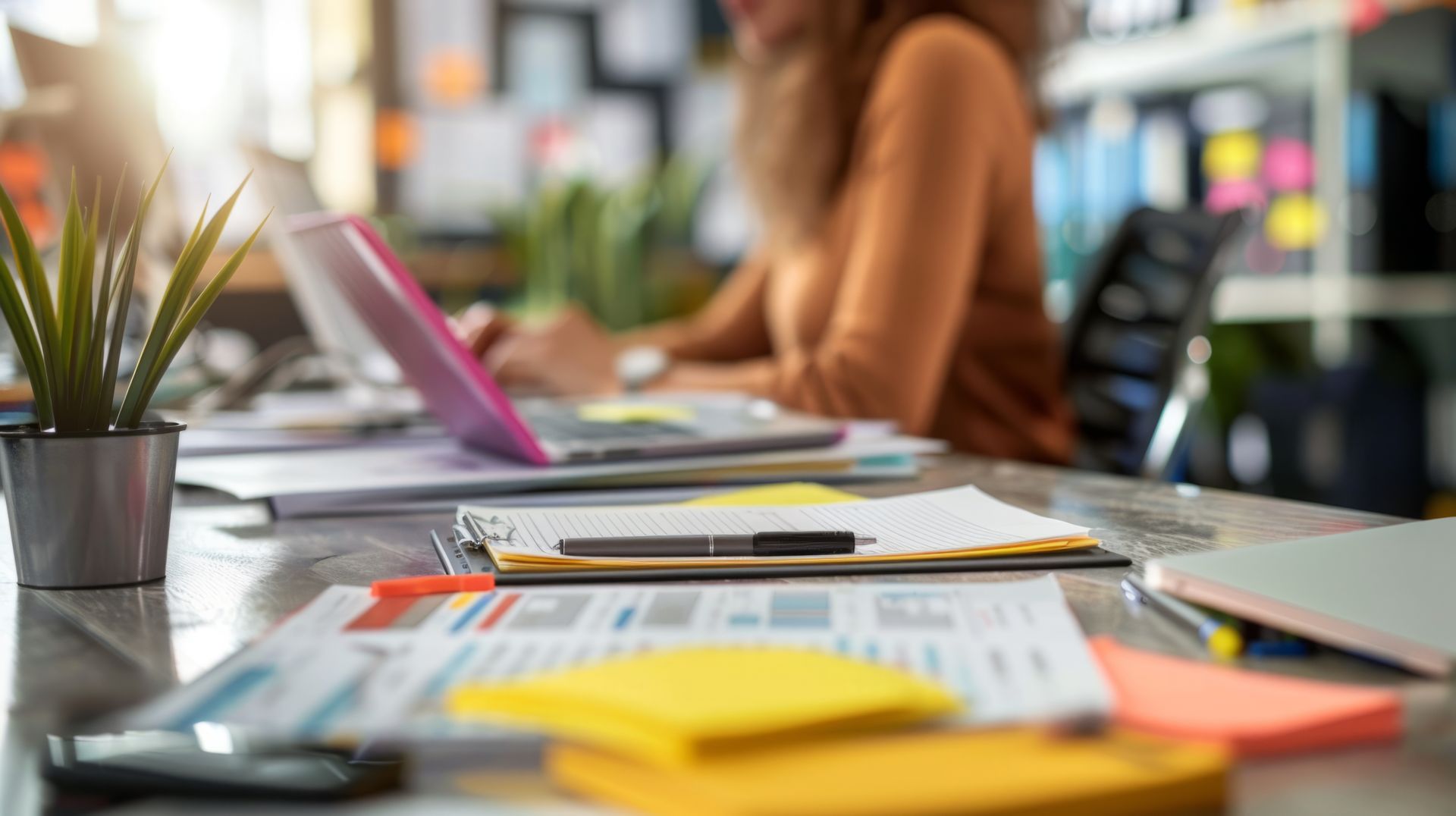 Une femme travaille sur un ordinateur portable à un bureau entouré de papiers, de notes adhésives et d'une plante en pot.