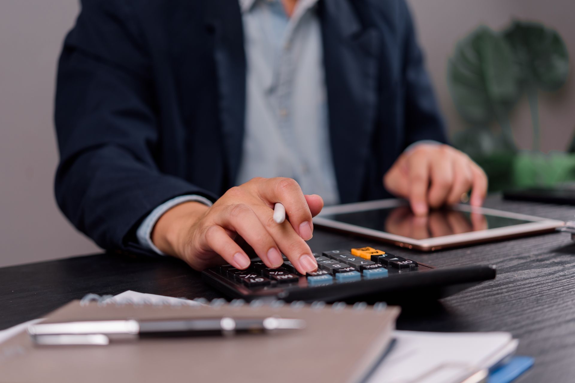Une personne en costume bleu utilise une calculatrice à un bureau, avec une tablette et un stylet.