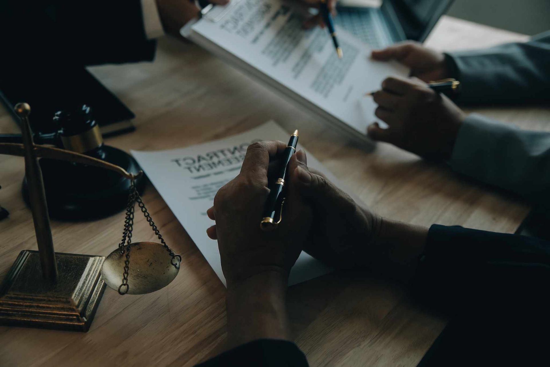 Persona firmando un contrato en un escritorio con una balanza de justicia, otras dos personas y documentos.
