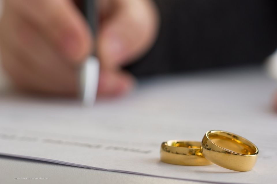 Person unterschreibt ein Dokument, auf dem Eheringe liegen