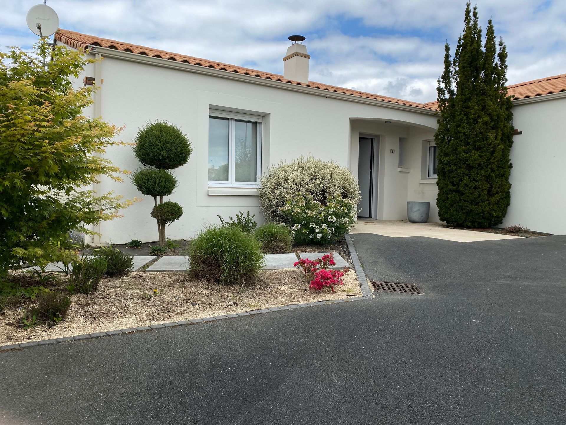 Une maison blanche avec un toit de tuiles, une allée de gravier et divers arbustes et arbres dans la cour avant.