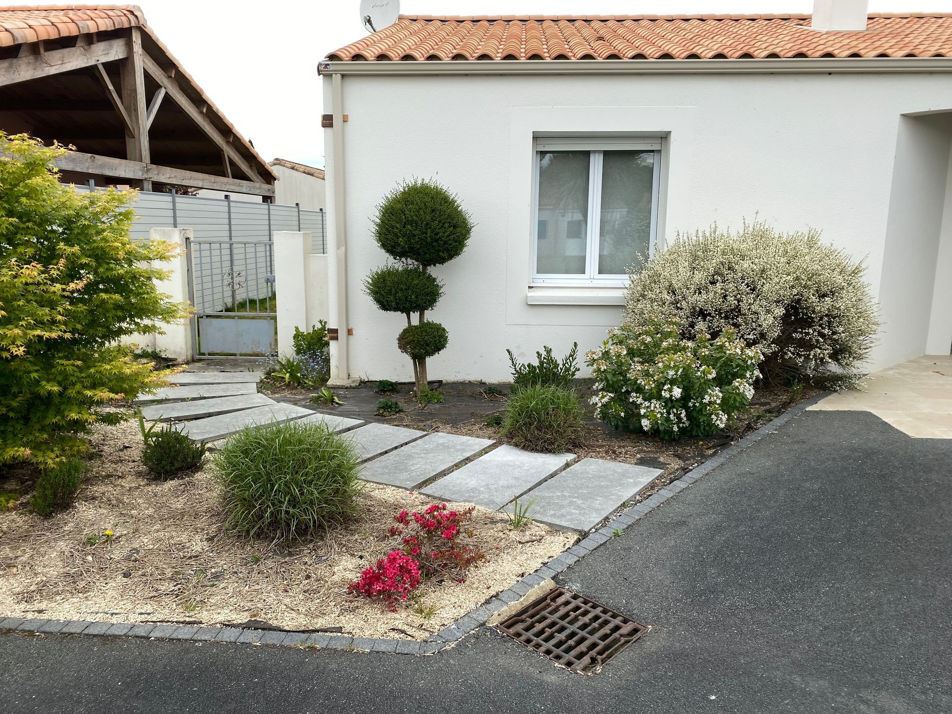 Un chemin de pierre traversant un jardin orné d'un arbuste taillé en topiaire, et un portail métallique gris à proximité.