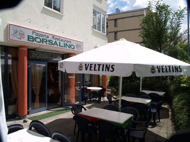 Sitzgelegenheiten im Freien mit weißen Veltins-Sonnenschirmen vor der Pizzeria Borsalino an einem sonnigen Tag