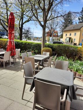 Außenterrasse eines Cafés mit Korbstühlen, grauen Tischen und einem roten Sonnenschirm neben einer Hecke und Gebäuden