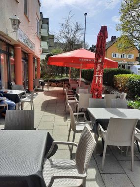 Außenterrasse eines Cafés mit grauen Tischen und Stühlen, roten Sonnenschirmen und einer von Gebäuden gesäumten Straße im Hintergrund