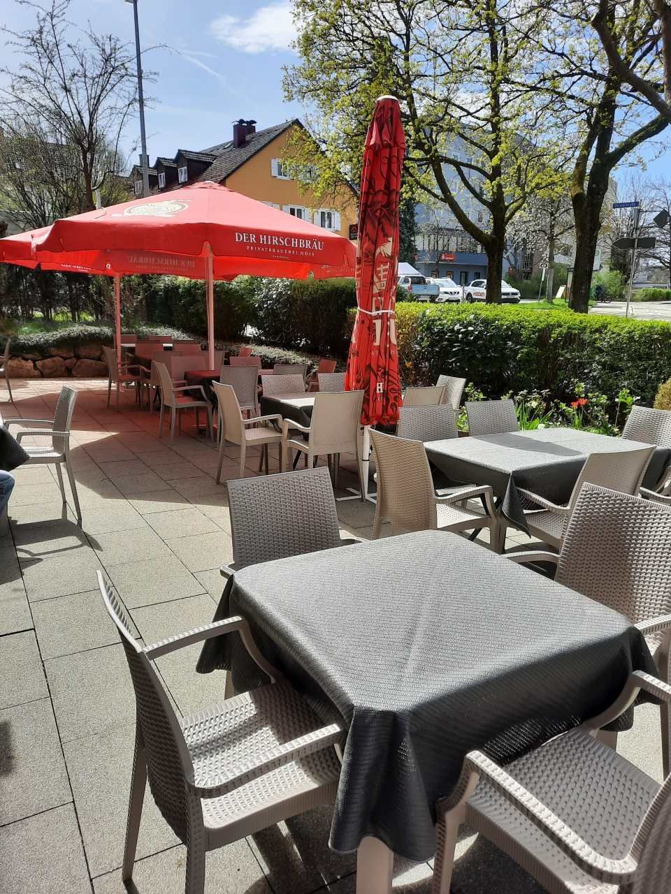 Terrasse eines Cafés im Freien mit Korbstühlen, Tischen und roten Sonnenschirmen bei sonnigem Wetter