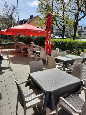 Terrasse eines Cafés im Freien mit Korbstühlen, Tischen und roten Sonnenschirmen bei sonnigem Wetter