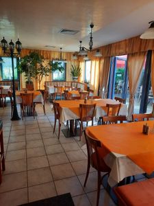 Leerer Speisesaal eines Restaurants mit orangefarbenen Tischdecken, Holzstühlen und großen Fenstern mit Blick ins Grüne