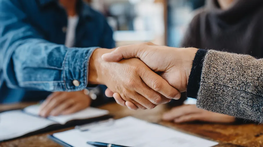Dos personas se dan la mano sobre unos documentos que están sobre una mesa, simbolizando un acuerdo.