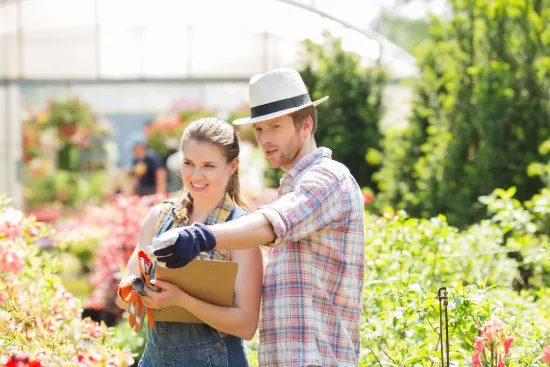 Un hombre y una mujer examinan plantas en un invernadero. El hombre señala, la mujer sostiene una tabla con notas; ambos visten ropa de trabajo.