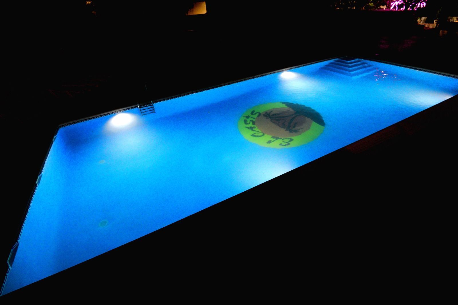 Una piscina rectangular por la noche, iluminada con luces azules brillantes, con un logotipo circular en el suelo.