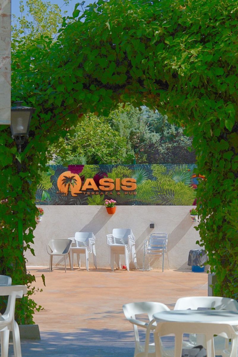 Un soleado patio al aire libre enmarcado por un frondoso arco verde, con sillas y mesas blancas y un letrero.