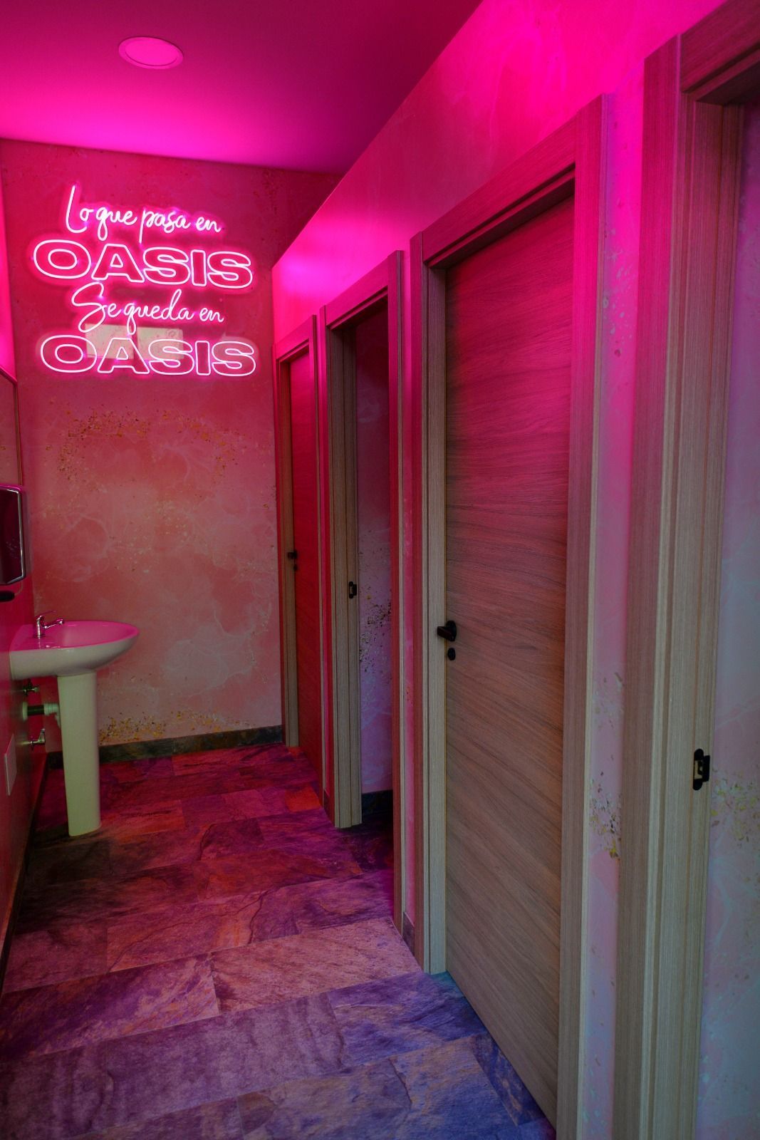 Un pasillo con iluminación de neón rosa, un suelo estampado, un lavabo y tres puertas con acabado de madera.