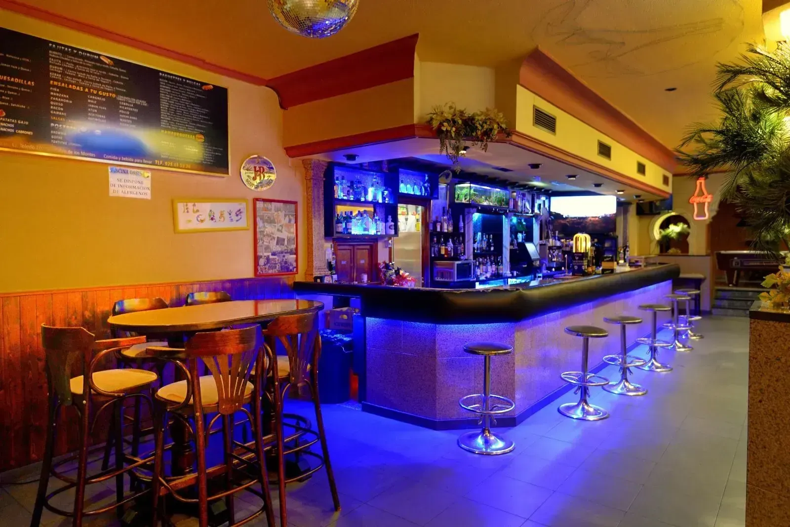El interior de un bar cuenta con una mesa de madera, varias sillas altas y una larga barra iluminada por luces .