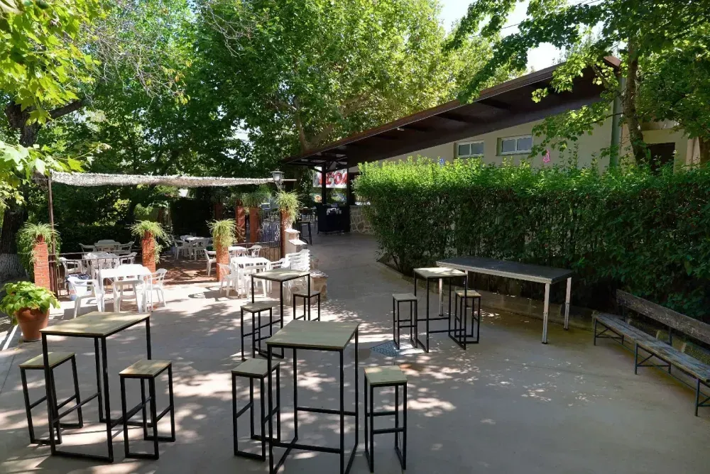 Un patio al aire libre con mesas altas, taburetes de bar y asientos informales bajo una pérgola sombreada.