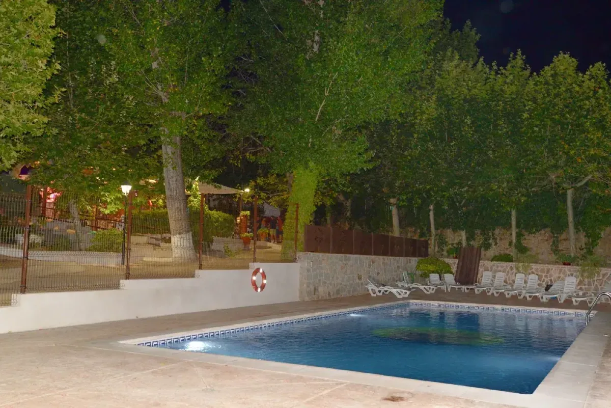 Una piscina al aire libre por la noche, rodeada por un muro de piedra, árboles y tumbonas bajo iluminación ambiental.