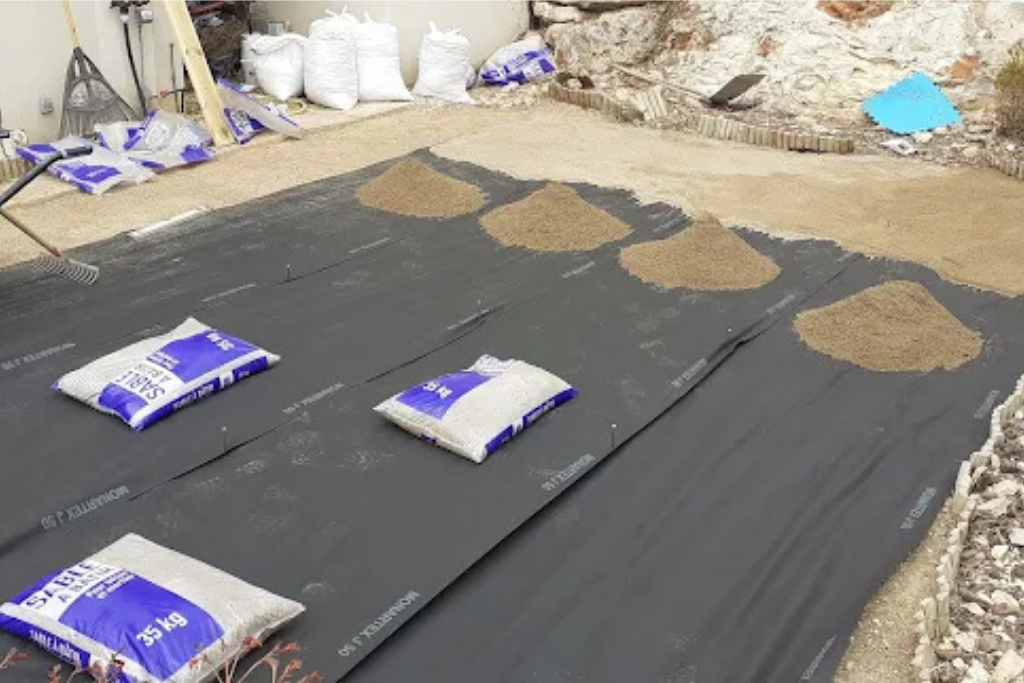 Tissu de paysage noir avec des sacs de tissu blanc et des tas de sable beige disposés dessus, dans un décor extérieur.