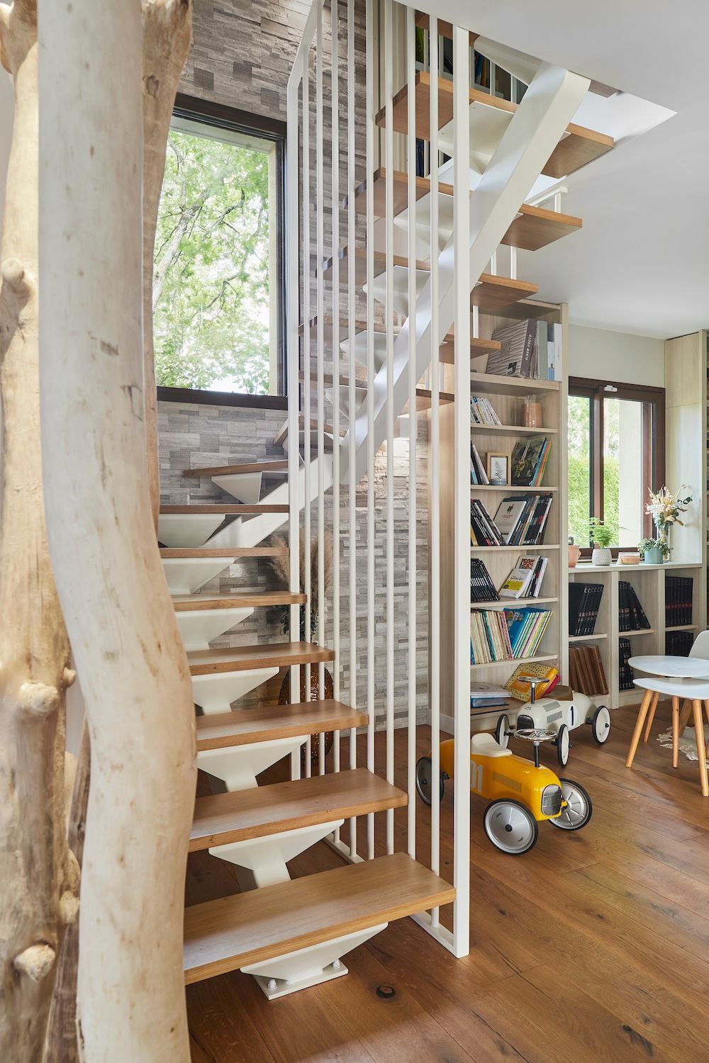 Intérieur moderne avec escalier en bois suspendu, rampes blanches et une pièce confortable avec étagères et fenêtres.