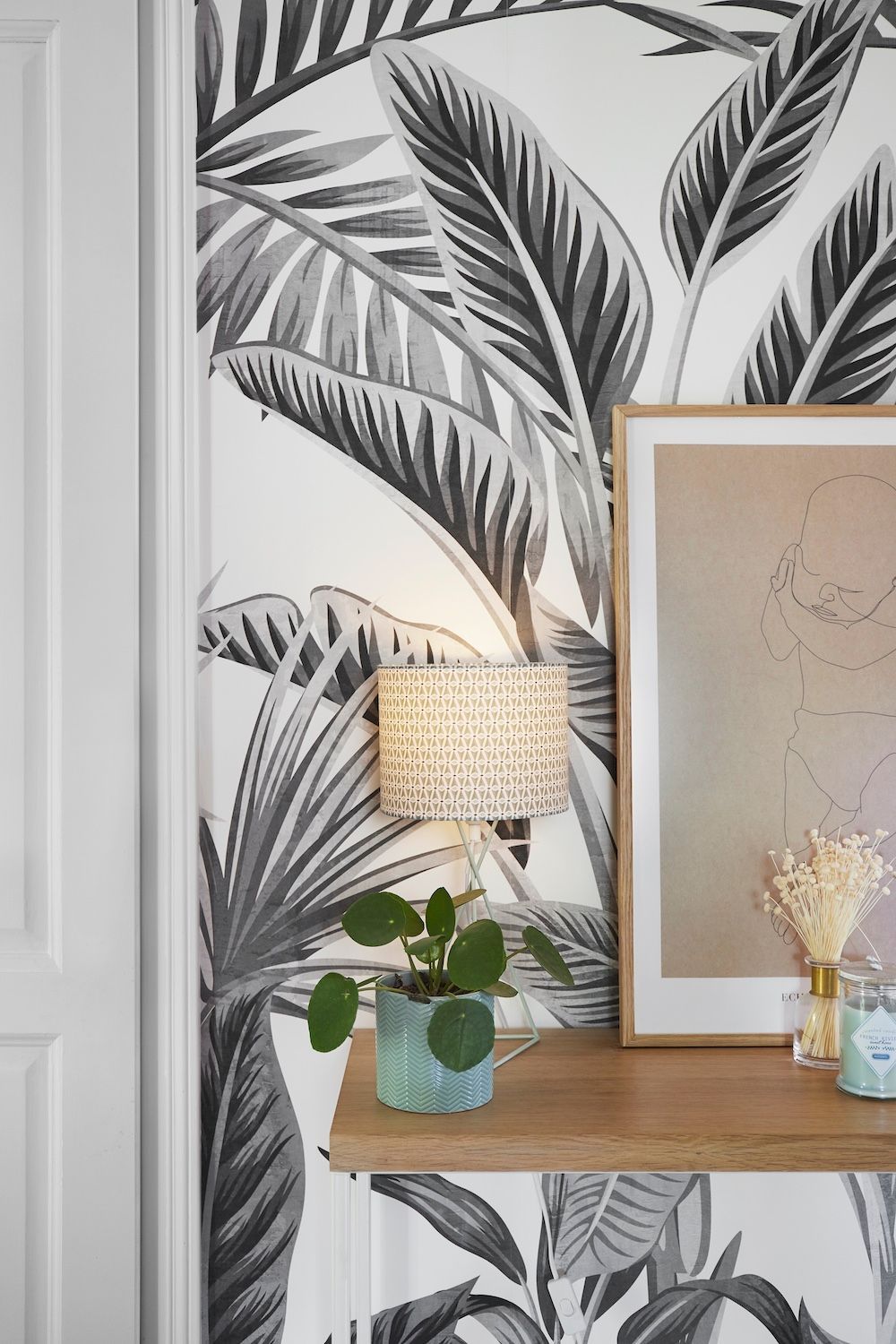 Papier peint à motifs de feuilles, console en bois, lampe, tableau encadré et plante en pot dans un coin lumineux.