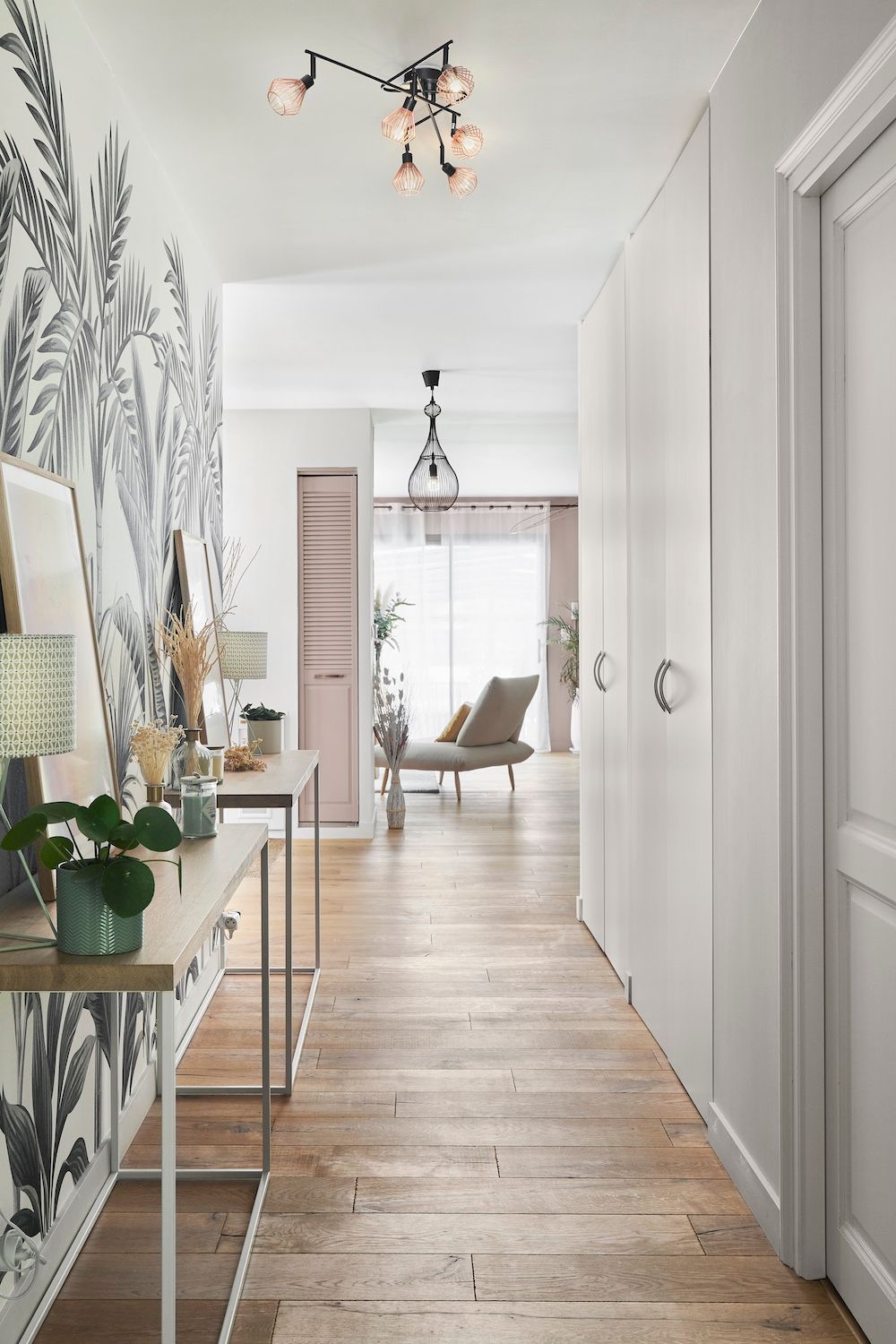Couloir lumineux avec murs blancs, parquet, papier peint fleuri et un salon visible au fond.