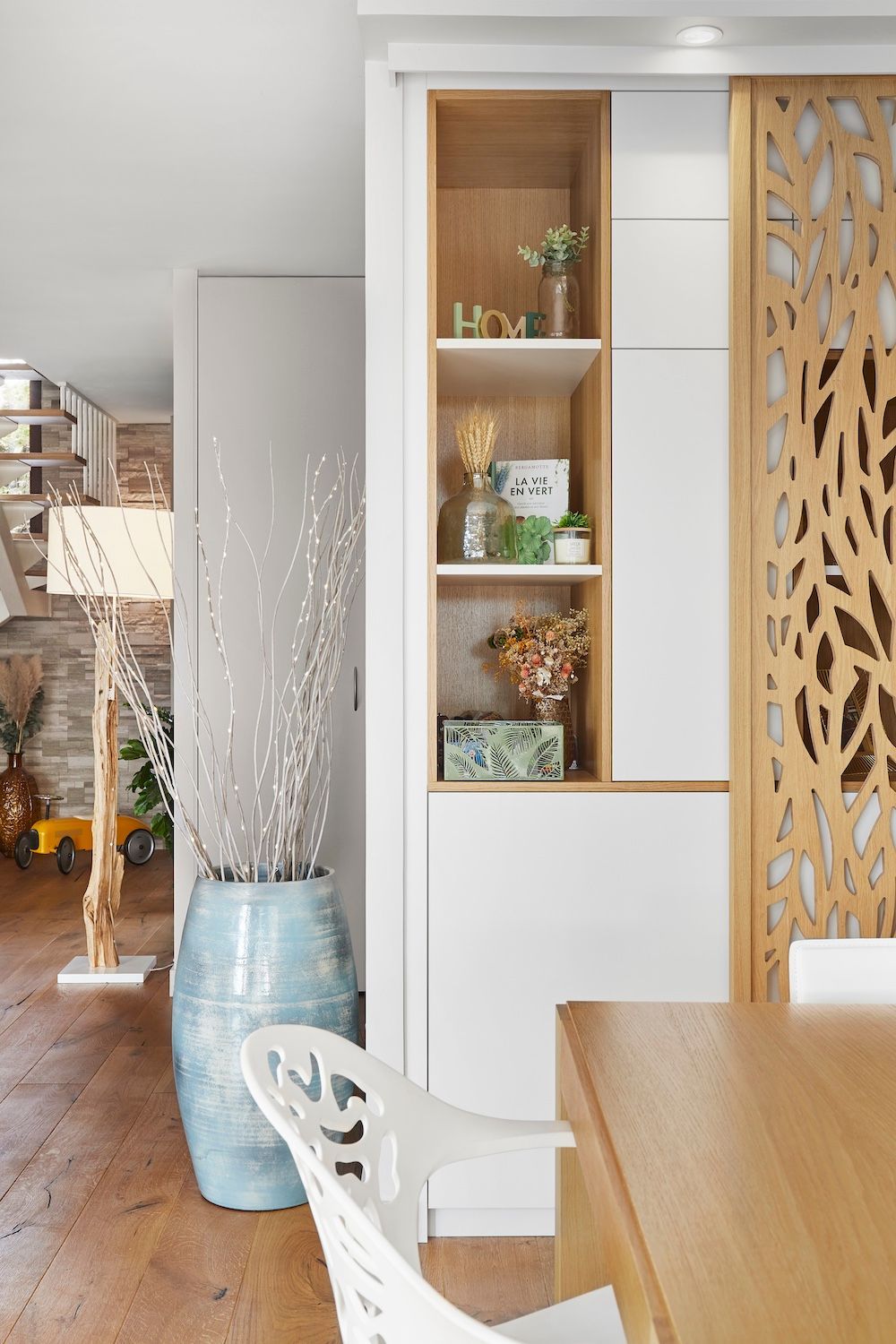 Salle à manger moderne avec étagères en bois, armoire blanche, grand vase bleu et paravent décoratif.
