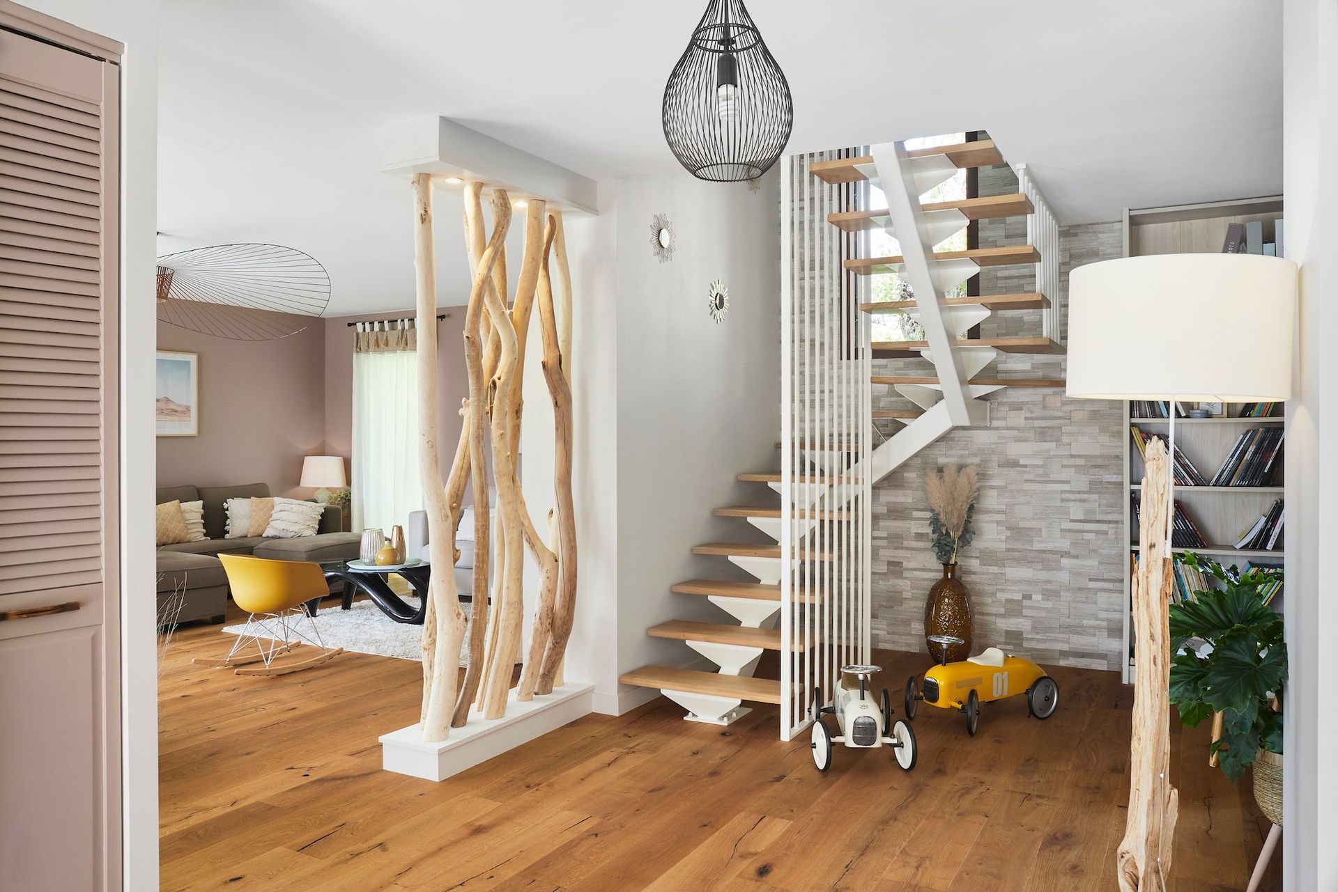 Salon moderne et lumineux avec escalier en bois, mur de briques blanches, suspension lumineuse et touches de jaune.