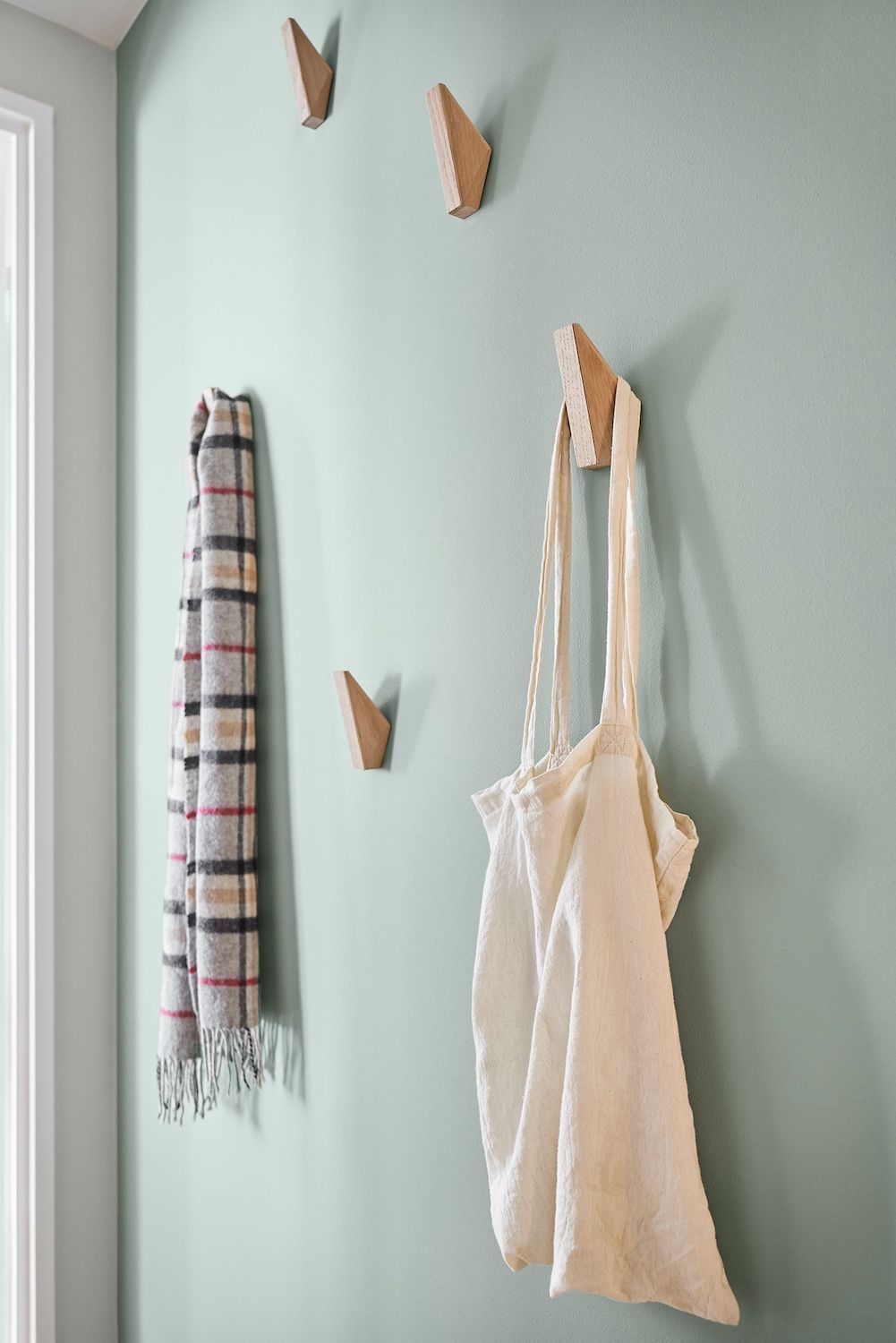 Des crochets muraux retiennent une écharpe rayée et un sac cabas crème contre un mur vert pâle.