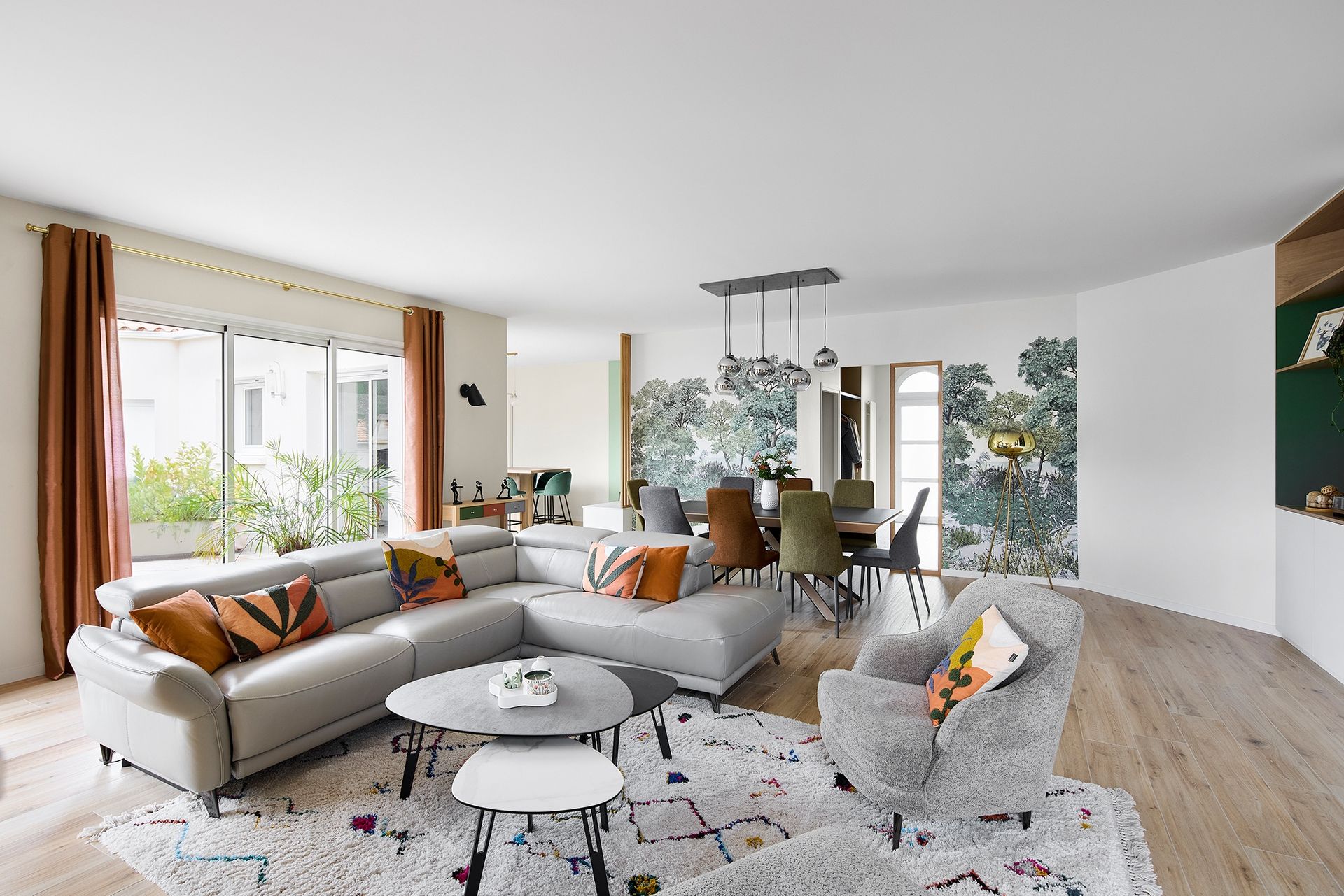 Salon lumineux avec canapé d'angle, fauteuils à motifs, coin repas et papier peint fleuri.