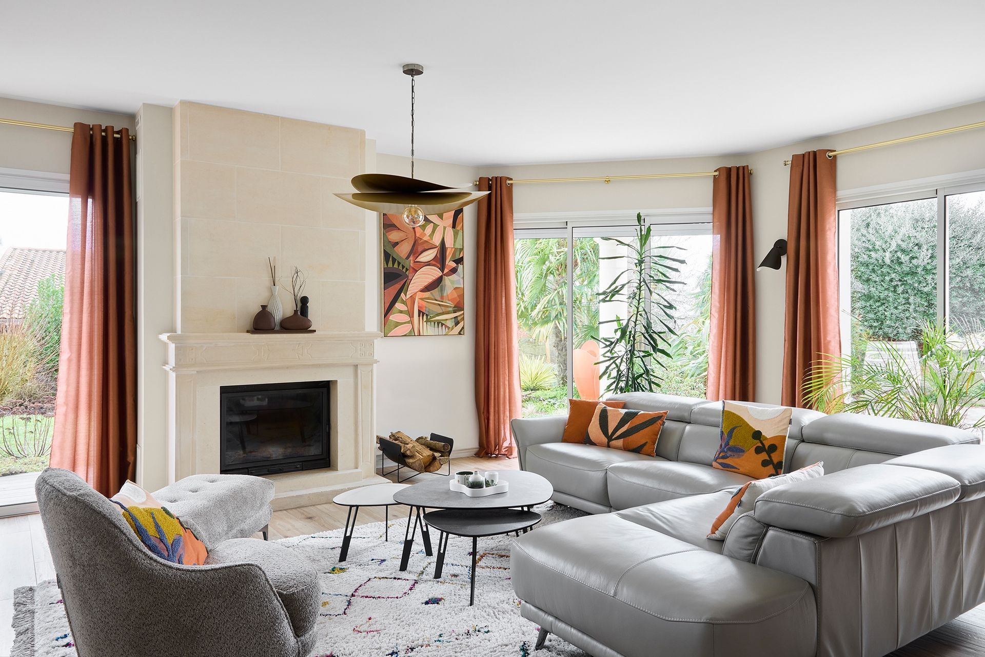Salon moderne et lumineux avec canapé d'angle gris, cheminée, grandes fenêtres et touches d'orange.