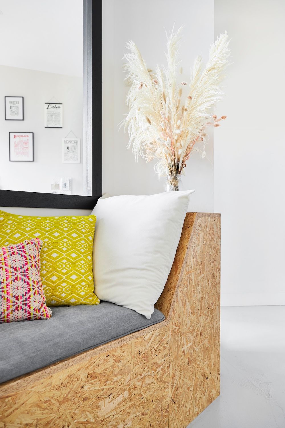 Canapé moderne avec coussin gris, oreiller à motifs jaunes et couverture blanche dans une pièce lumineuse avec un vase d'herbes séchées.