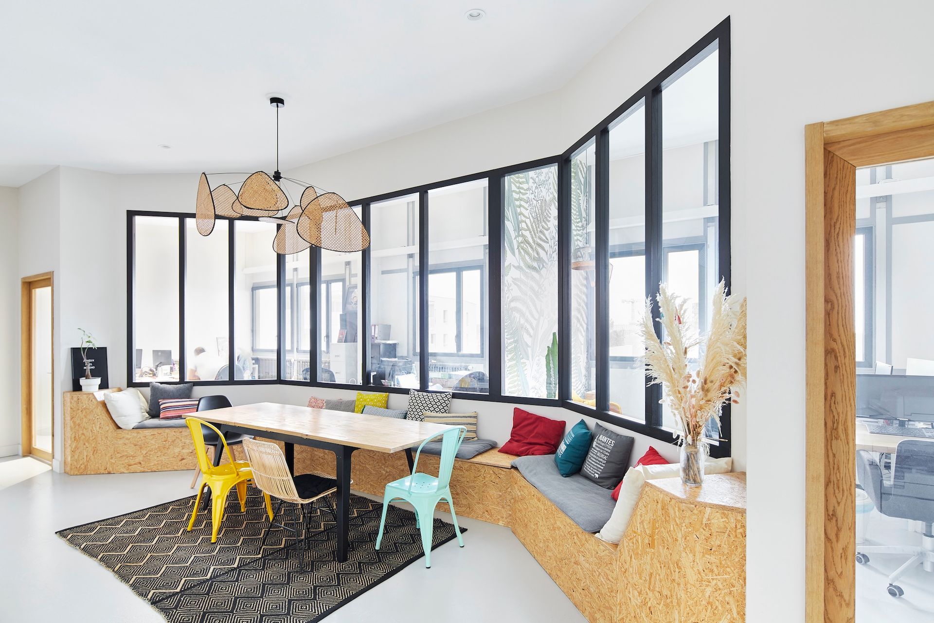 Salon moderne et lumineux avec fenêtres à cadre noir, banquette incurvée, chaises jaunes et tapis noir