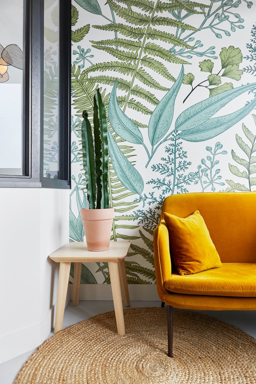Un canapé jaune moutarde à côté d'une petite table en bois et d'une plante en pot devant un mur tapissé de papier peint à motifs de feuillage.