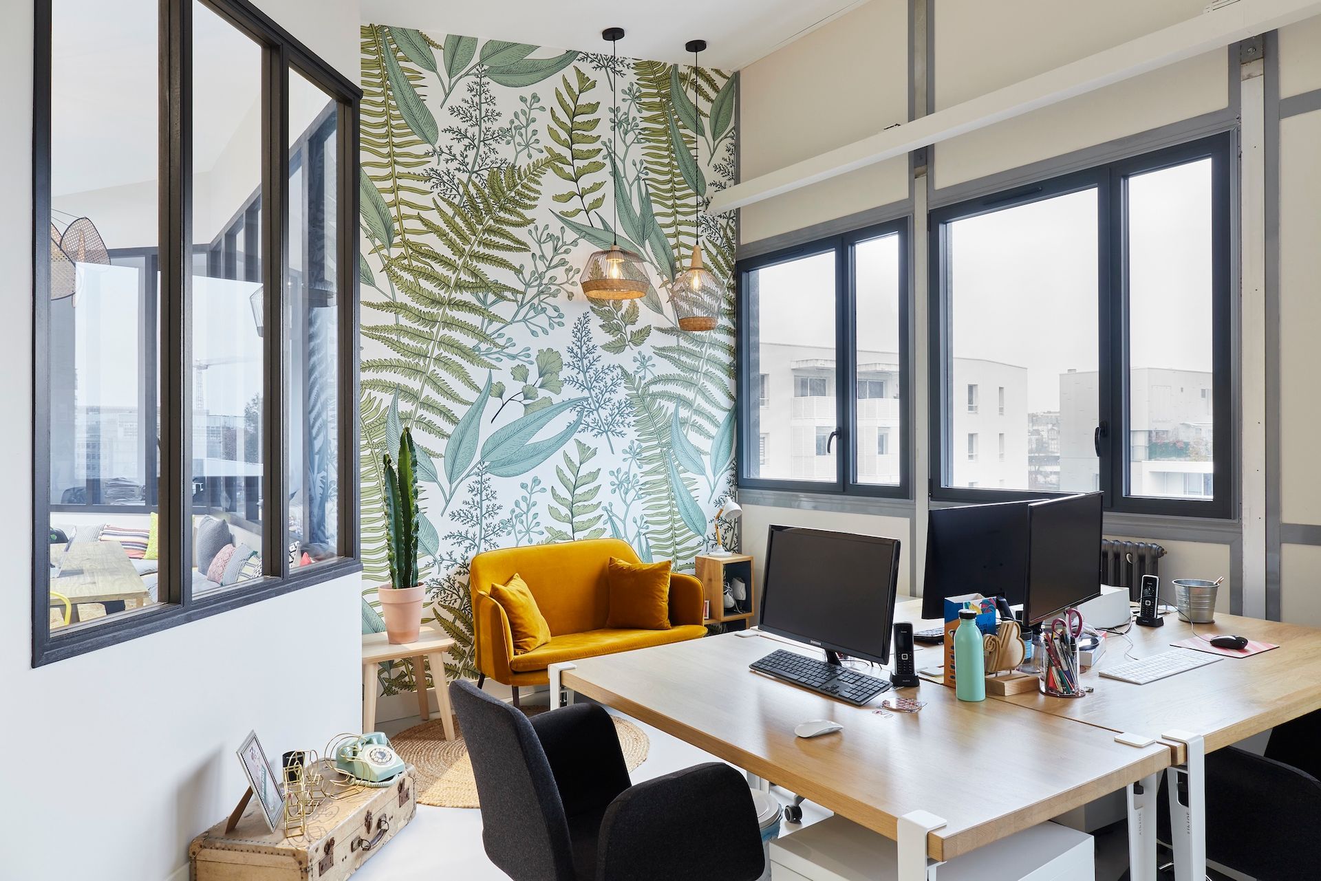 Bureau moderne avec bureaux, chaises noires, canapé moutarde et décoration murale à motifs végétaux près de grandes fenêtres