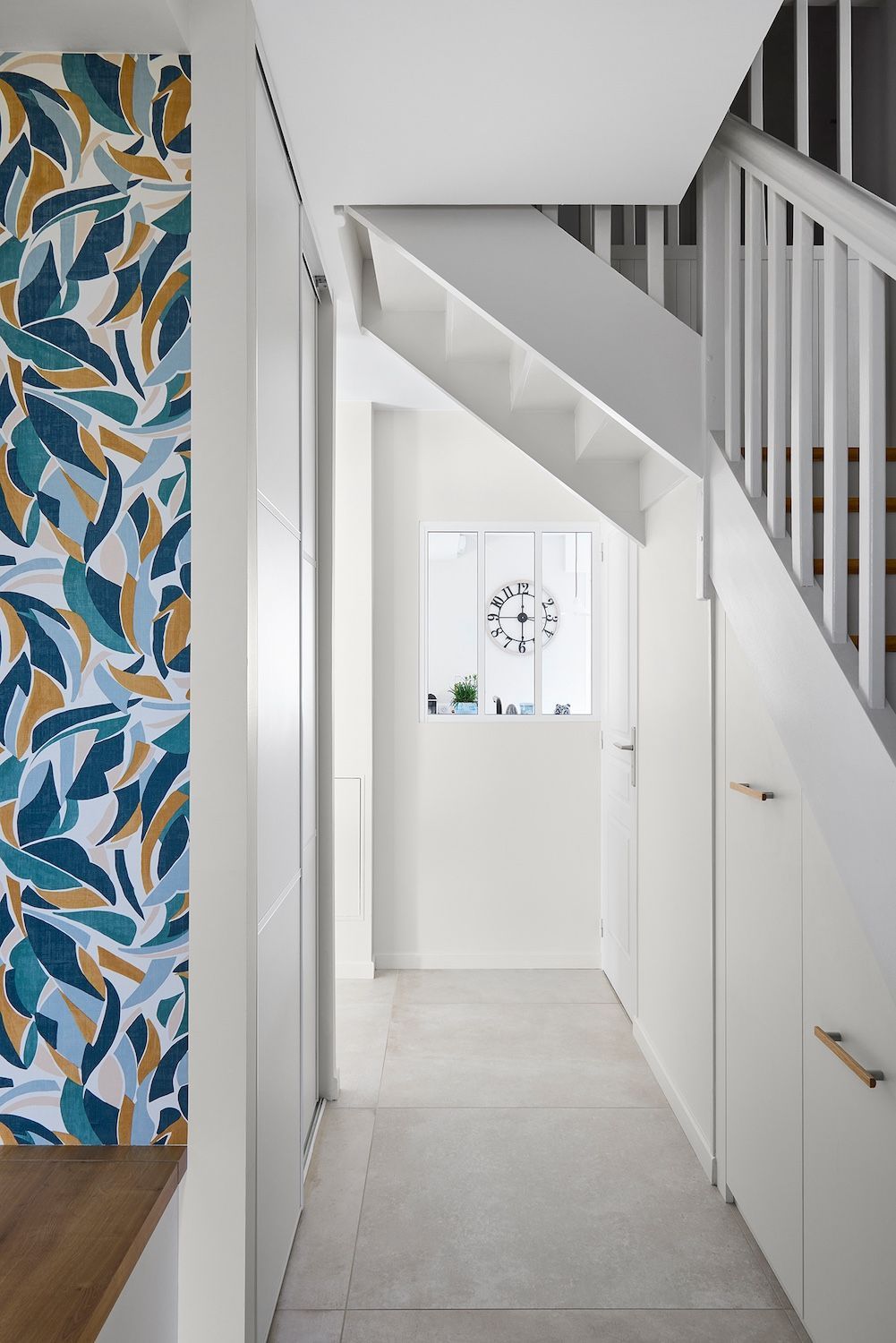 Couloir étroit et blanc avec escalier, sol carrelé et papier peint à motifs sur la gauche.