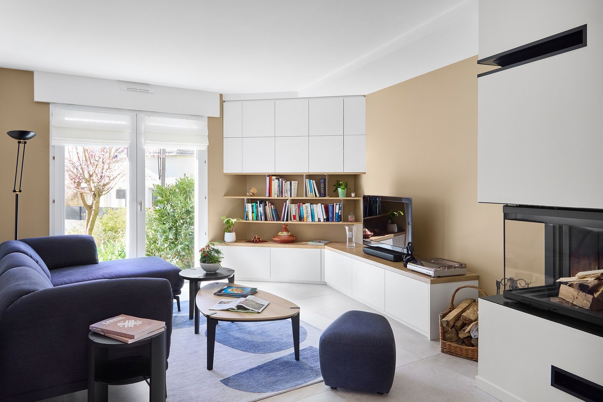 Salon moderne et lumineux avec canapé bleu, meubles blancs, télévision et grande fenêtre donnant sur le jardin.