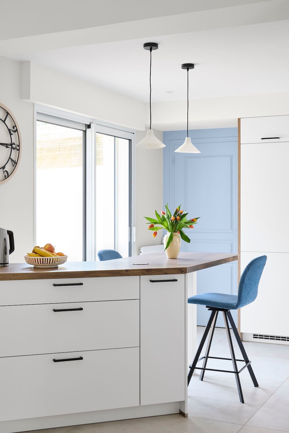Cuisine moderne et lumineuse avec placards blancs, îlot en bois, tabouret de bar bleu et fleurs sur le comptoir