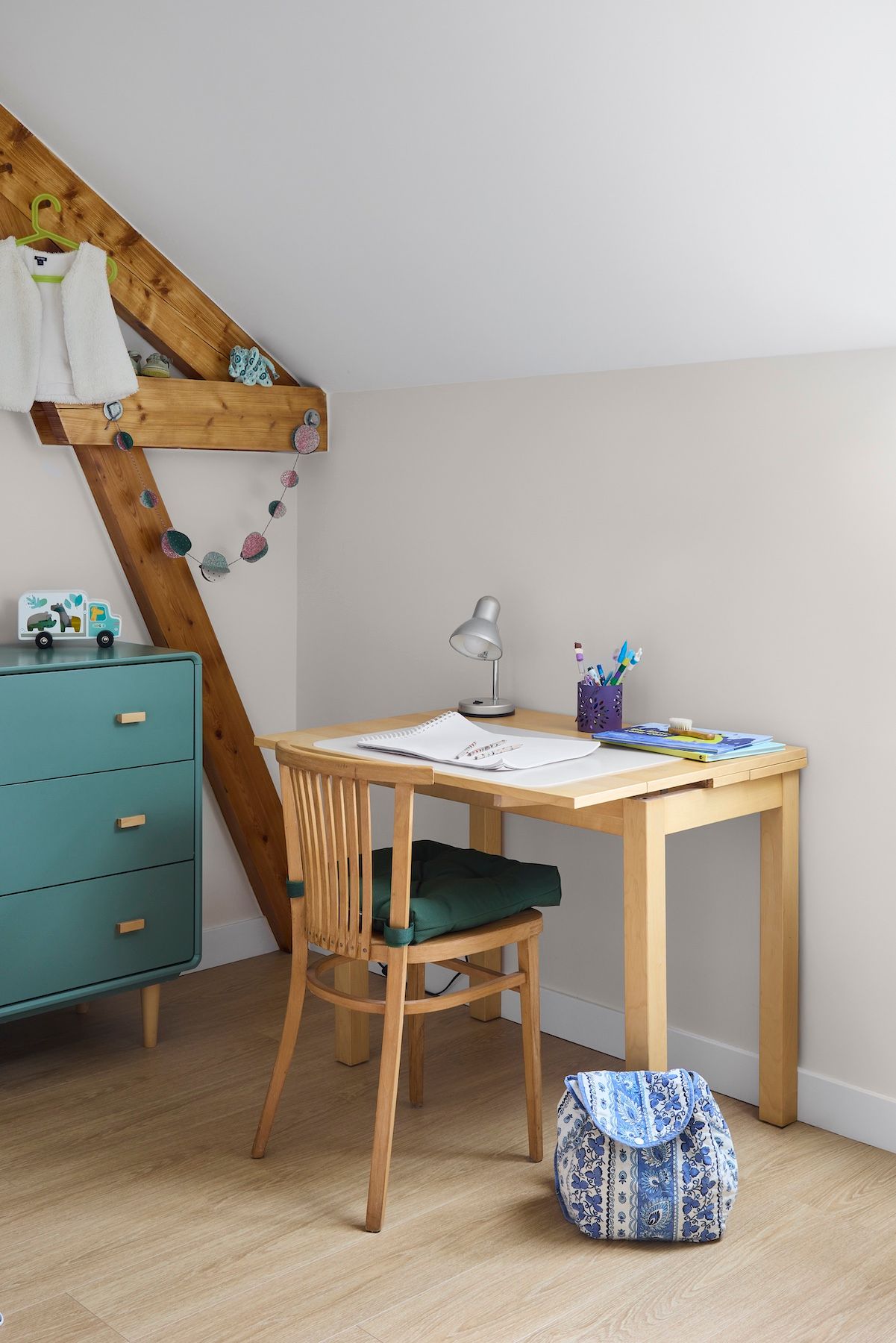 Petite chambre mansardée avec un bureau en bois, une chaise, une commode turquoise et un sac à motifs posé au sol.