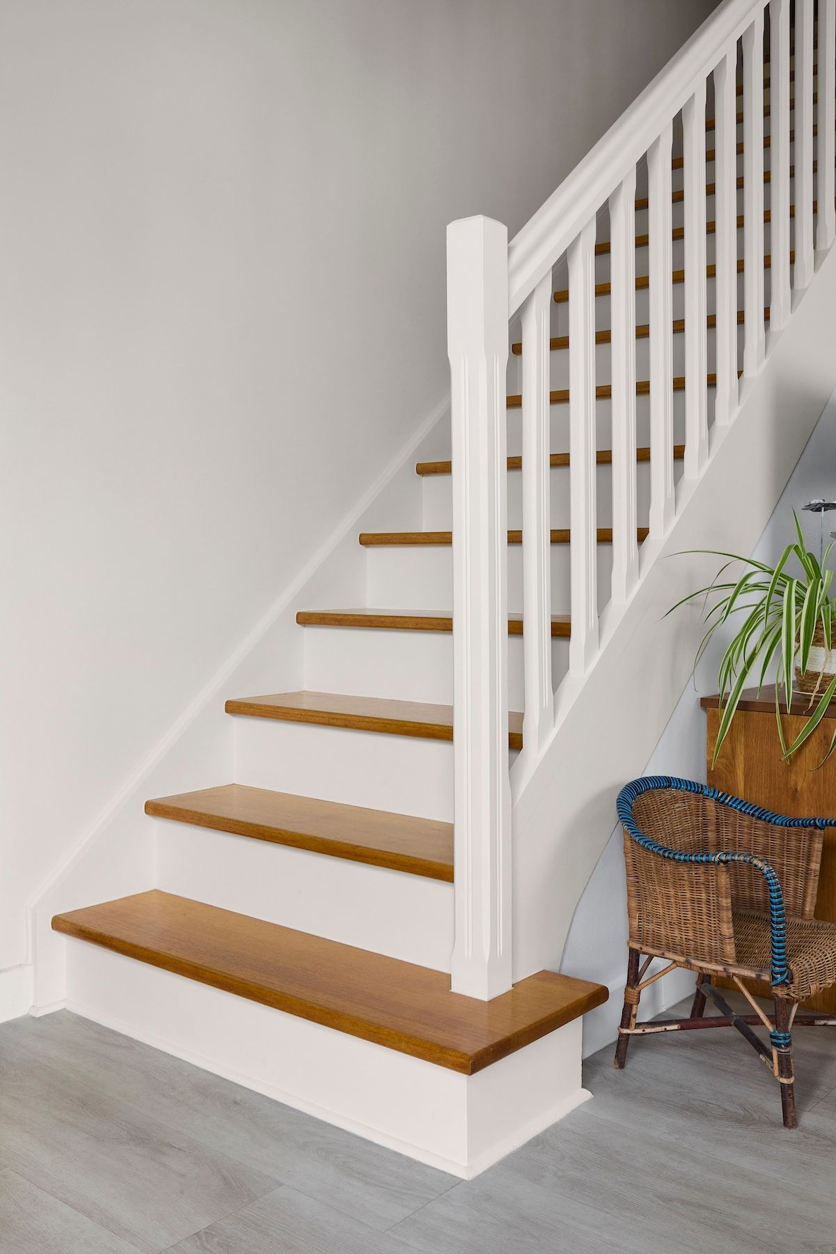 Un escalier blanc avec des marches en bois, une rampe blanche et une plante à côté d'un fauteuil en osier dans un intérieur de maison