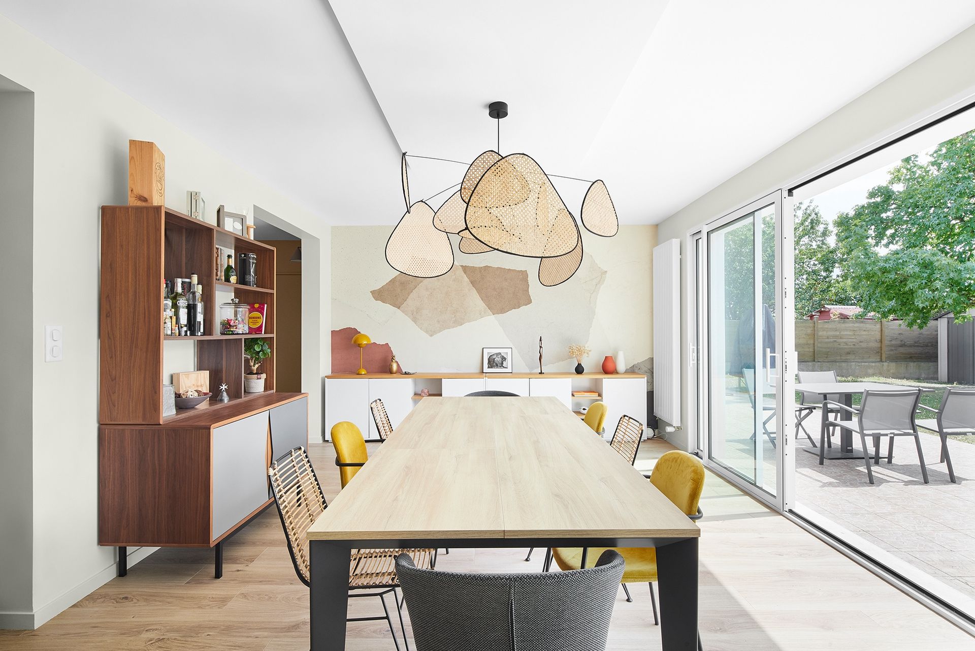 Salle à manger lumineuse avec table en bois, suspensions lumineuses en fibres naturelles et portes coulissantes en verre donnant sur une terrasse