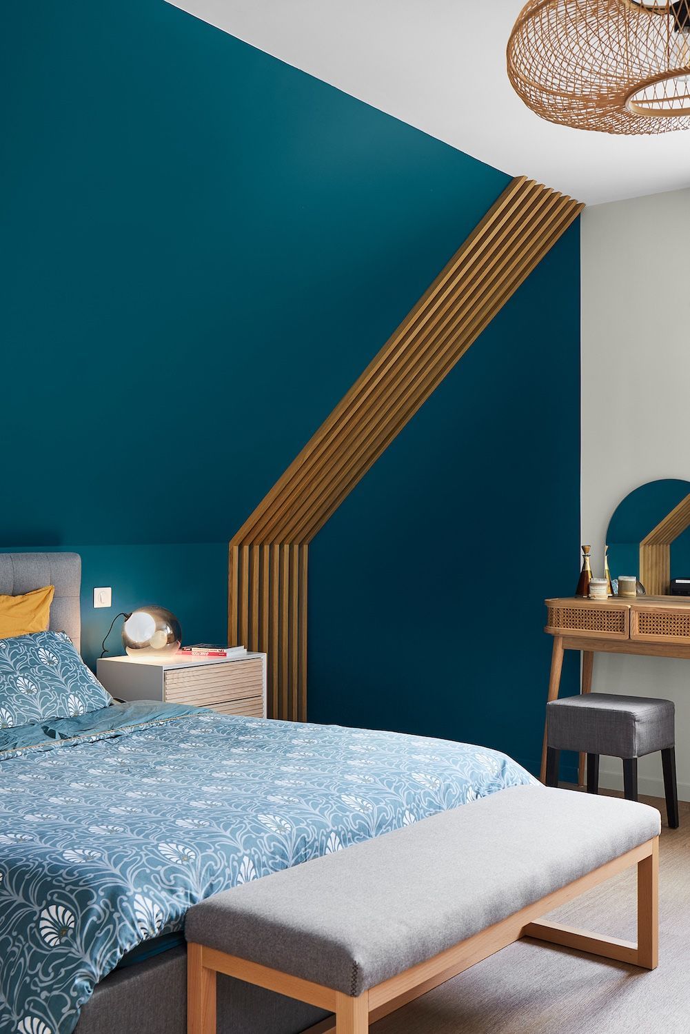 Chambre bleu sarcelle avec accents de bois inclinés, lit, banc, table de chevet et miroir de coiffeuse.