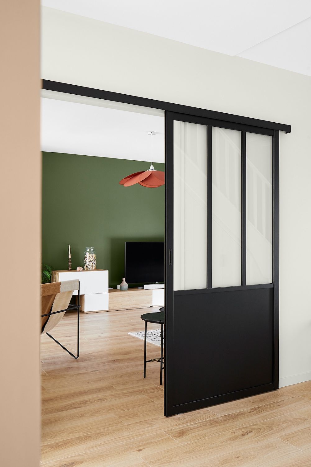 Porte coulissante en verre à cadre noir donnant sur une pièce avec mur vert, parquet et chaises