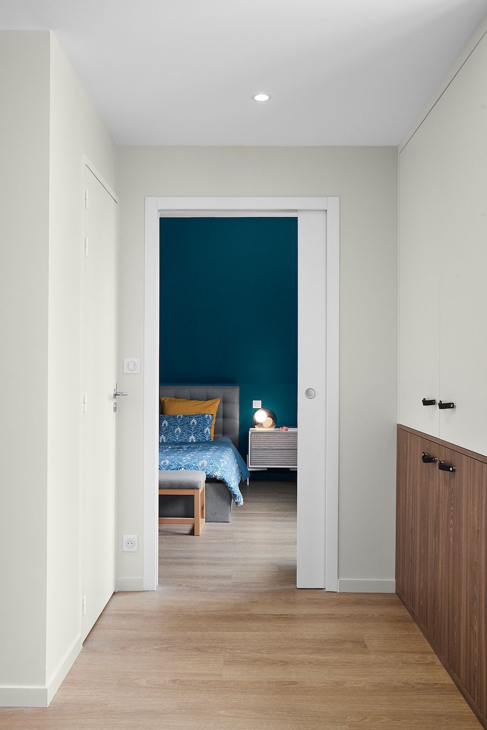 Couloir avec parquet clair menant à une chambre avec un mur d'accent turquoise et un lit visible à travers la porte.