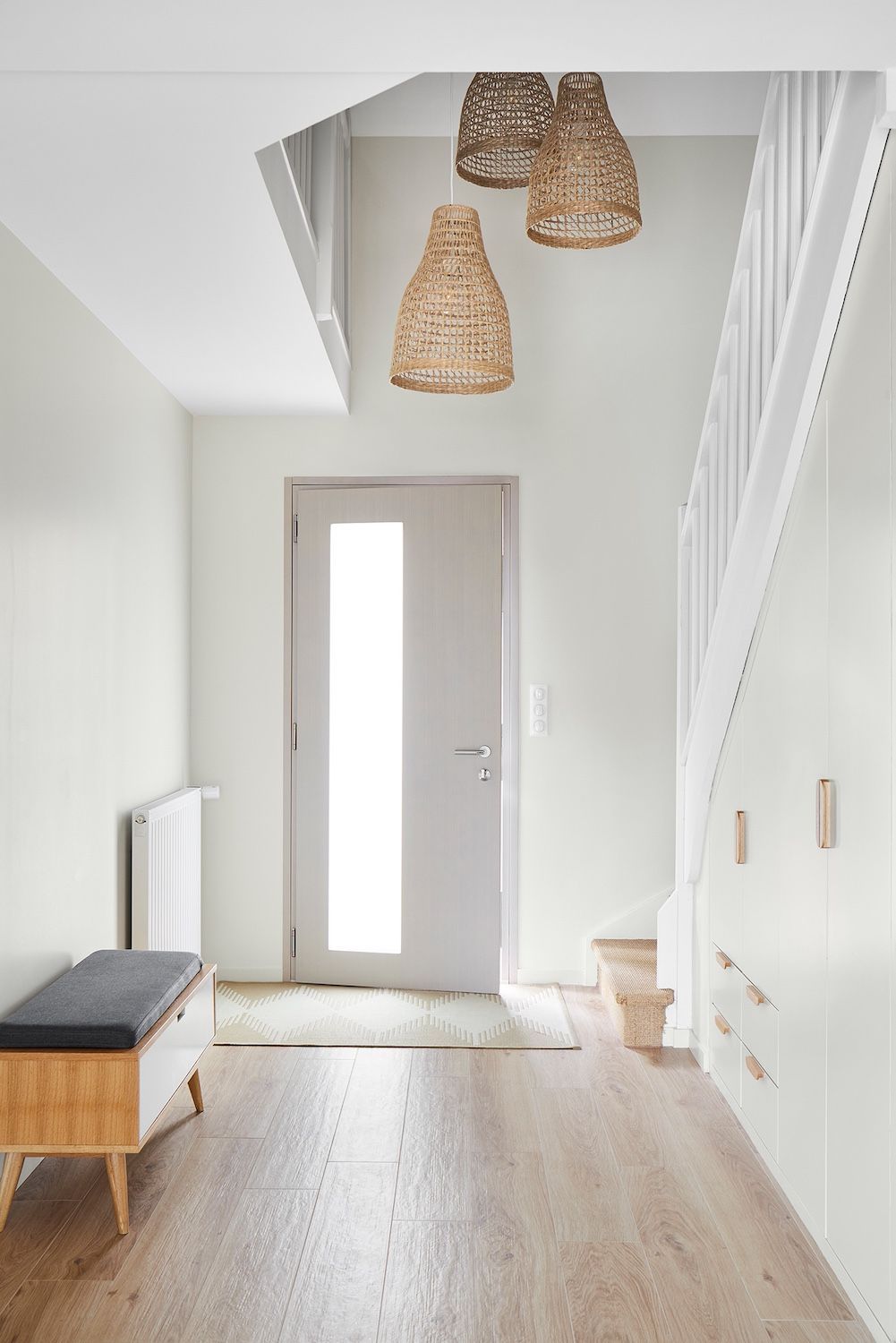 Couloir lumineux avec parquet, porte d'entrée grise, suspensions lumineuses en tissu tressé et rampe d'escalier blanche.
